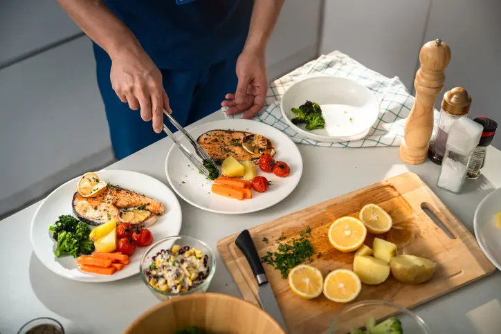 Person plating salmon with vegetables and lemon.